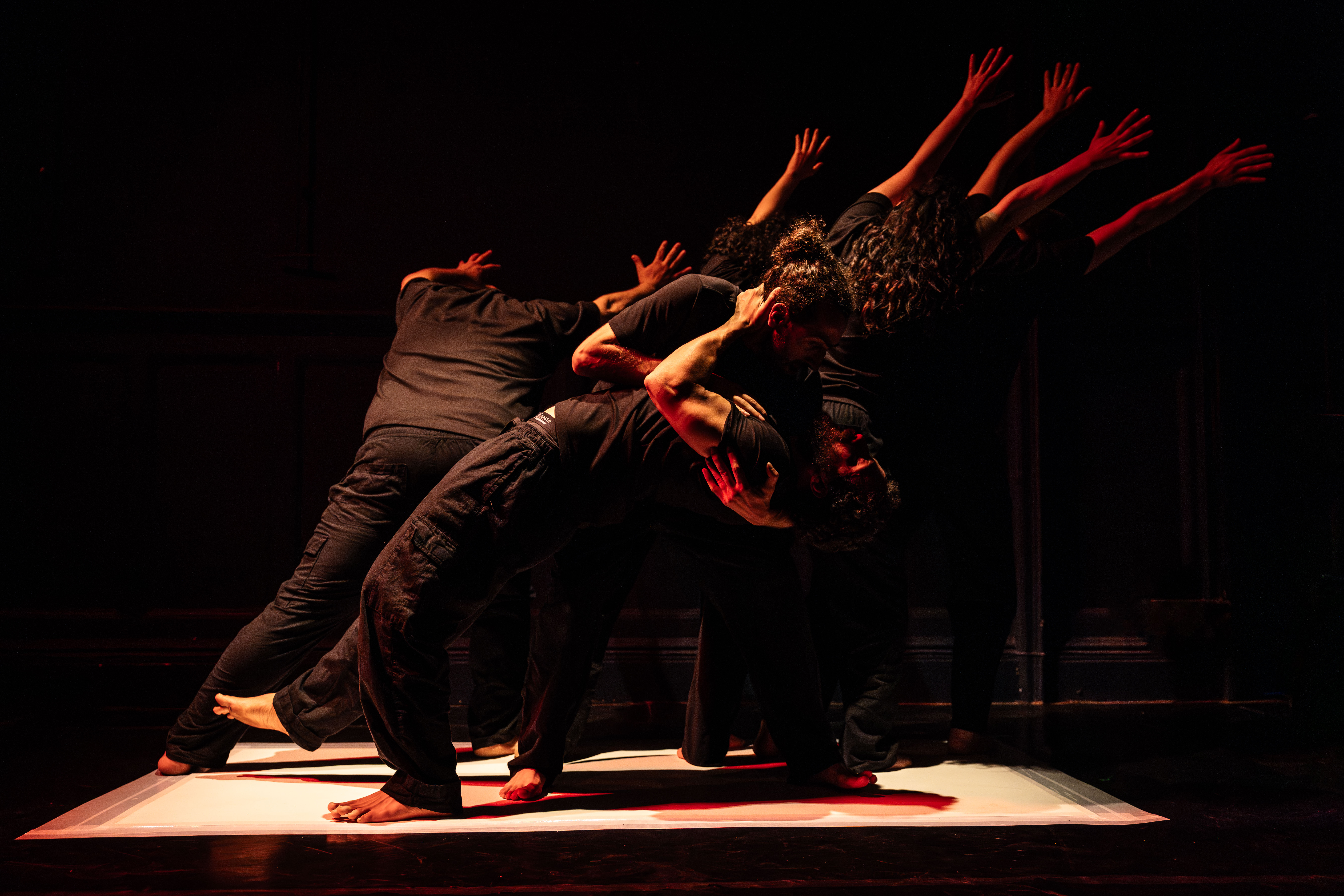 A group of performers dressed in black moves together in a powerful contemporary dance formation on a dimly lit stage. Their bodies lean and reach in unison, creating a sculptural shape under warm red and amber lighting, conveying strength, resistance, and collective movement.