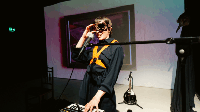 Image Desciption: A production photo from ‘The King of All Birds’ showing Performer Martha Knight smiling, and playing a vocoder. Behind her there is an old painting projected on the wall and a mandolin. She is wearing a black jumpsuit with orange straps, and there are flight goggles over her eyes.