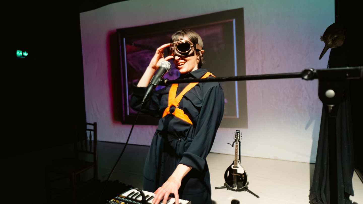 Image Desciption: A production photo from ‘The King of All Birds’ showing Performer Martha Knight smiling, and playing a vocoder. Behind her there is an old painting projected on the wall and a mandolin. She is wearing a black jumpsuit with orange straps, and there are flight goggles over her eyes.