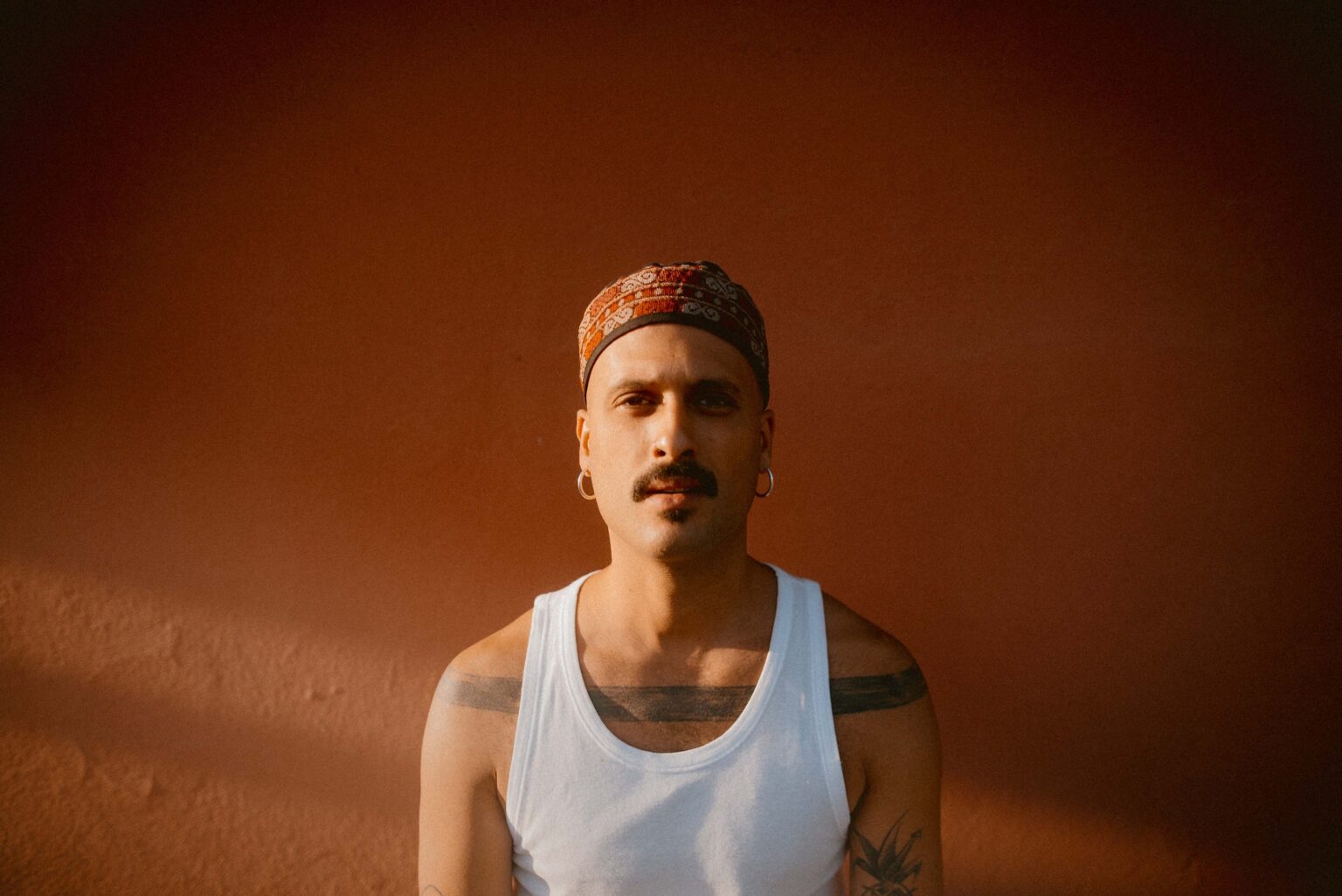 Musician Ahmed Eid looks into the camera. The background is a deep burnt orange wall. He is wearing a white vest, a tattoo band clearly visible across his chest from each shoulder. From each ear hangs silver hoop earrings. He is wearing a deep red and orange hat with a Tatreez design.