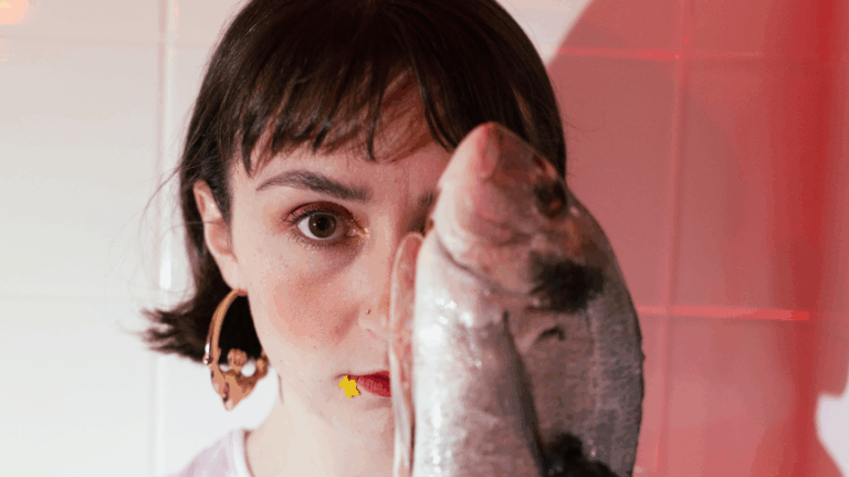 A young white woman with short dark hair stands against a white tile background holding up a dead fish to cover half of her face as she looks directly at the camera. Tá bean óg gheall le gruaig ghearr dhorcha ag seasamh in aghaidh balla le tíleanna bána sa chúlra, le greim aici ar iasc marbh a chlúdaíonn leath dá haghaidh, agus í ag breathnú i dtreo an cheamara.